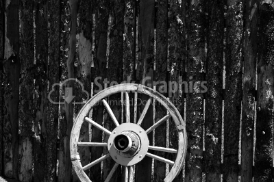 An old rusty wagon wheel leaning on a barn wall in black and whi