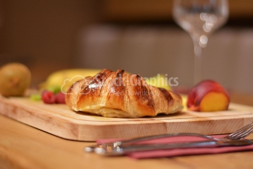 Croissant with various fruits around, placed on a wooden platter.