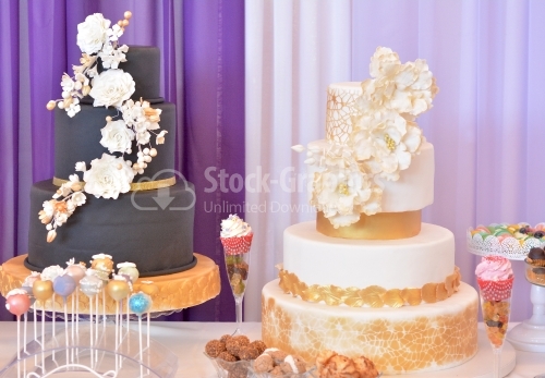 Two big cakes. cake covered with black marzipan, golden ribbon and beige flowers. White cake with golden decorations, large beige flowers, golden lace and golden leaves
